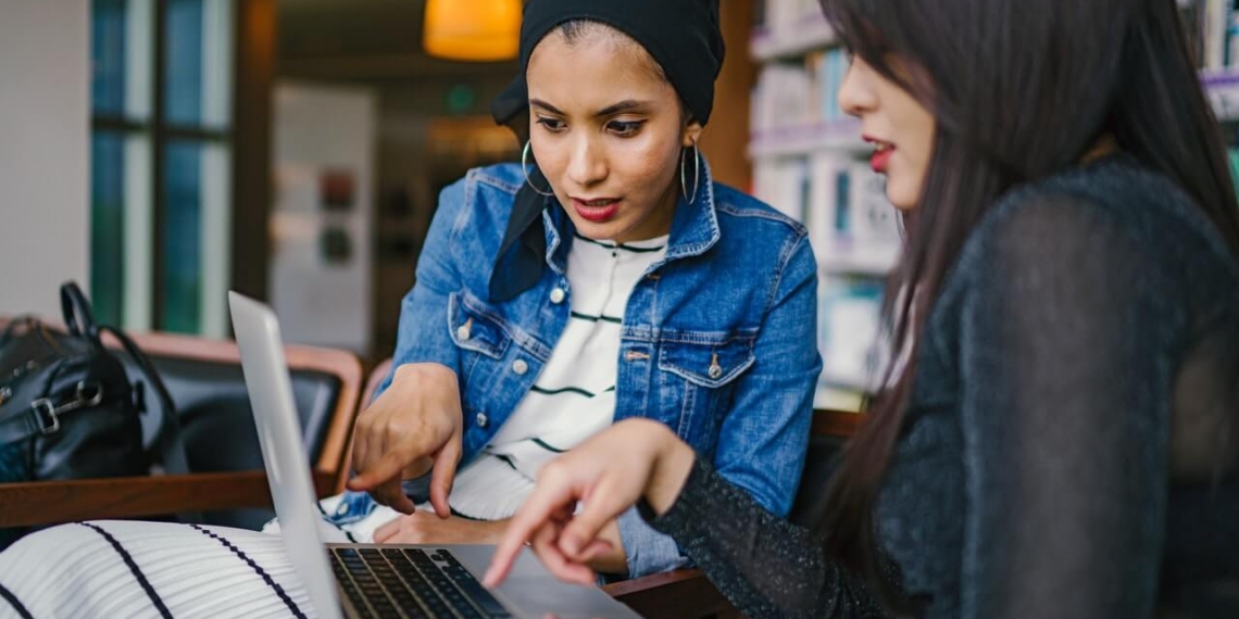 Two muslim women looking at a laptop in a library, engaged in discussions regarding business news.
