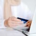 A woman holding a credit card in front of a laptop while browsing business news.