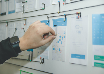 A person is pointing at a board filled with business news and a lot of paper.
